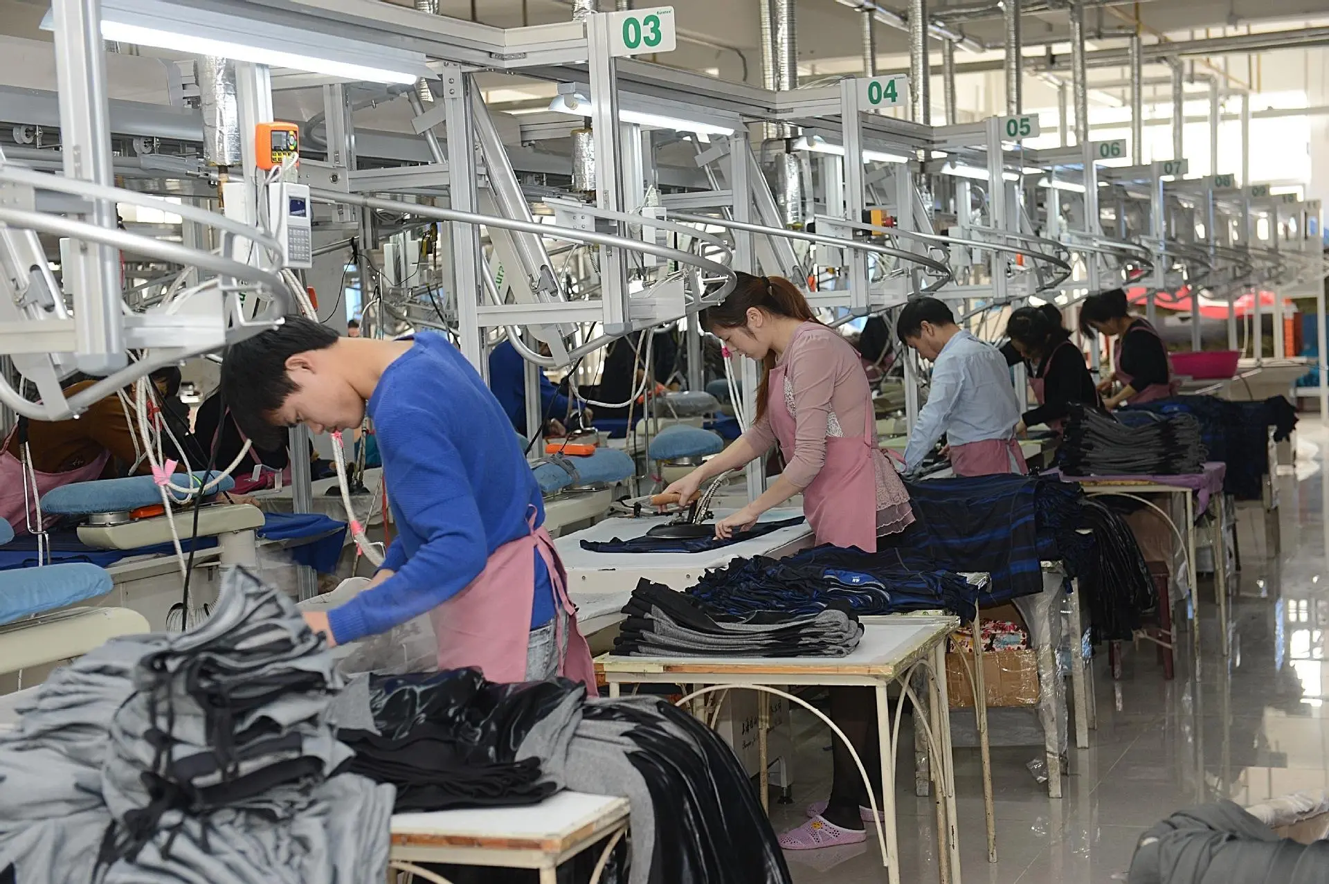 This image captures a corner of the Aprilluck yoga wear manufacturing workshop, showcasing the precision and craftsmanship that goes into every piece. The perspective of the image is from a medium angle, highlighting the various machines and workstations that occupy the space. The composition is well-balanced, with the machines and workers arranged in an orderly manner. The lighting is bright and natural, emphasizing the clean and organized nature of the workspace. The colors are primarily white and neutral, creating a professional and modern atmosphere. The style of the image is documentary, with no obvious filters or effects applied.